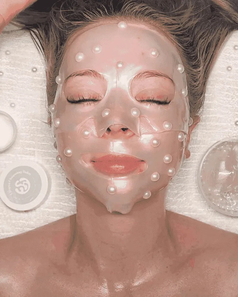 A guest resting with eyes closed under a pearl-studded jelly mask, facial products beside her
