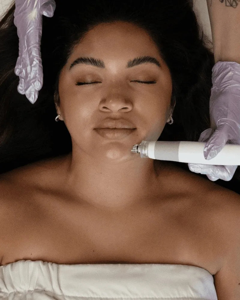 An esthetician in lavender gloves guiding a Hydrafacial handpiece along a guest's jaw